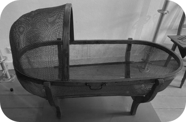 Black-and-white photograph of a historic metal structure with a rectangular frame and wire mesh, presumably an early industrial or forge product.